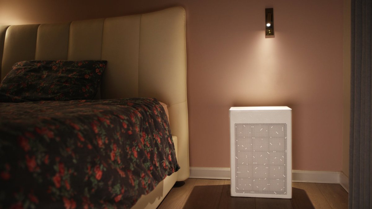 Smart Air SA600 air purifier with bedside lamp shining on it next to a bed with pink wall in background