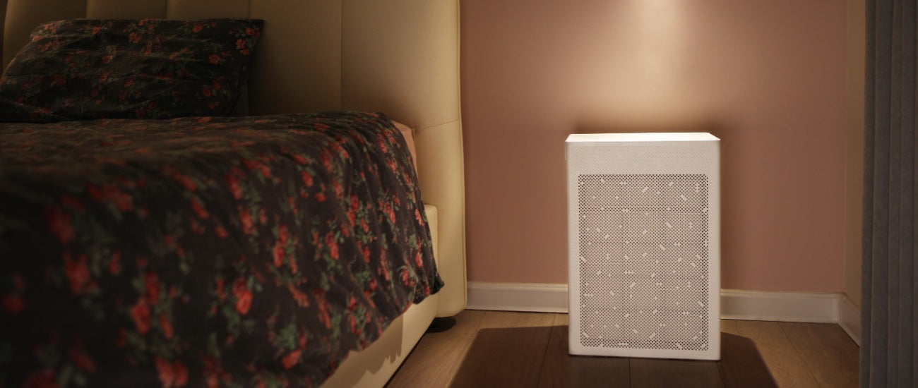 Smart Air SA600 air purifier with bedside lamp shining on it next to a bed with pink wall in background
