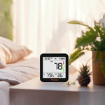 Temtop M10+ displaying AQI index reading plus temperature and humidity on a table with a plant and pillows in the background