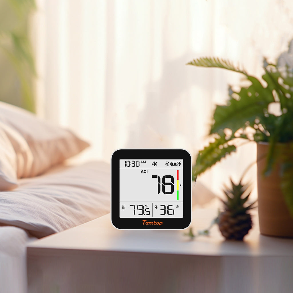 Temtop M10+ displaying AQI index reading plus temperature and humidity on a table with a plant and pillows in the background
