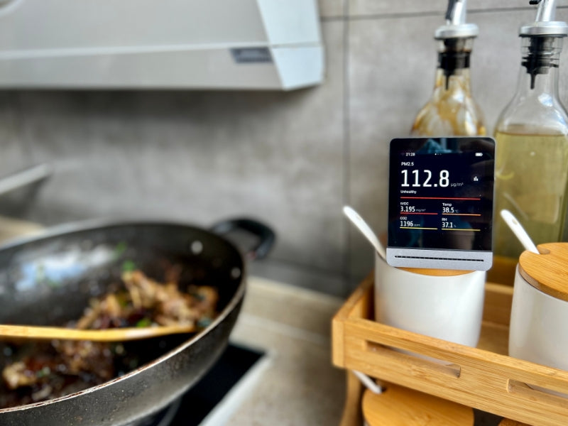 Qingping Pro air quality monitor displaying high PM2.5 and TVOC readings in a kitchen setting with cooking pan and ingredients.
