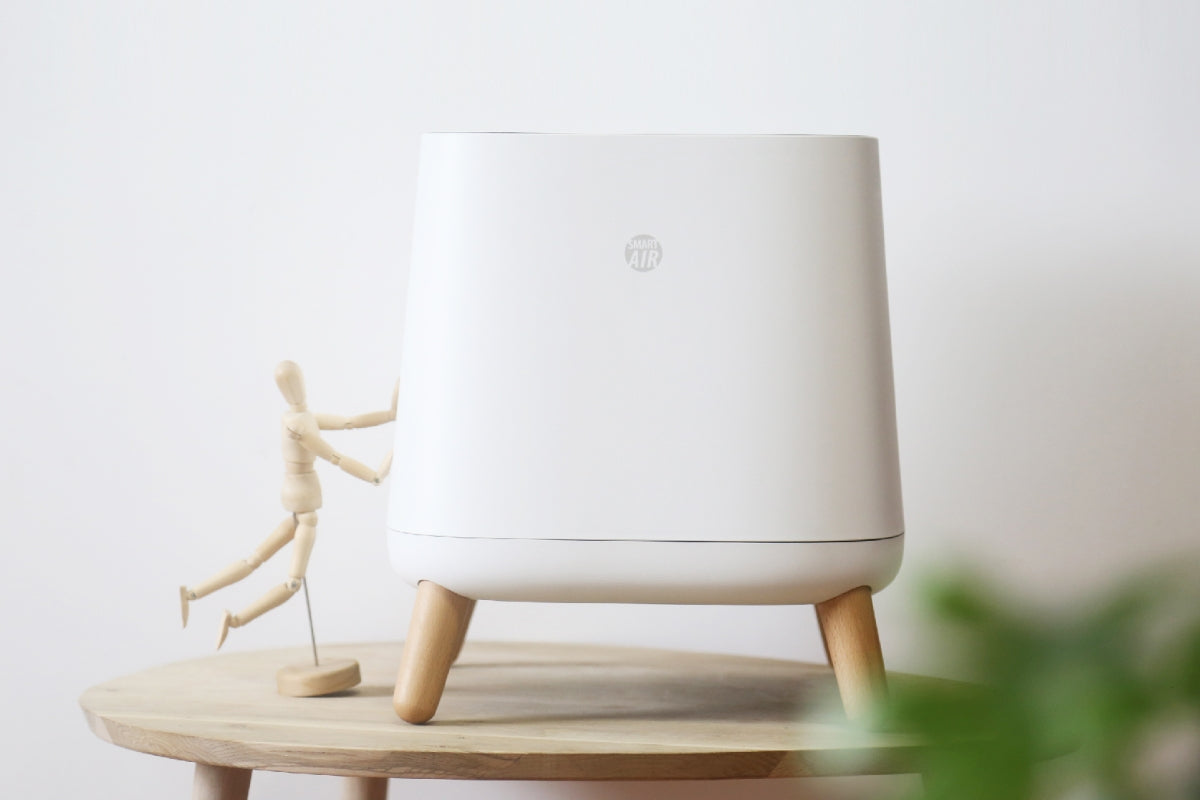Small wooden figure pushing a Smart Air Sqair air purifier on a wooden table with a white wall in the background