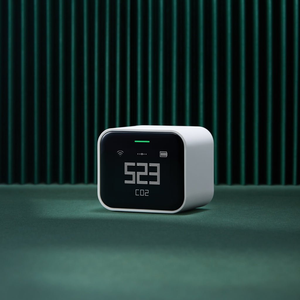 QP Lite Air Quality Monitor on a green surface with dark green vertical slat wall in background